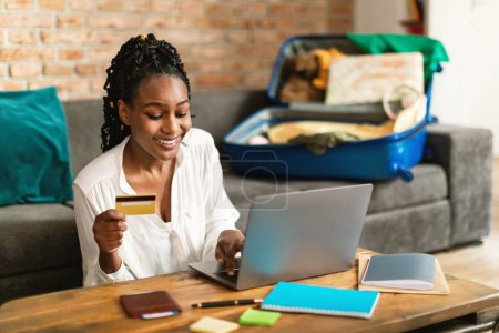 Téléchargez les photos : Heureuse femme noire utilisant un ordinateur portable et réservation tour en ligne avec carte de crédit, assis avec valise déballée sur le canapé à la maison, se préparer pour les vacances, espace libre - en image libre de droit