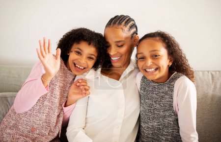 Photo for Hi. Cheerful African American Mom And Two Daughters Posing Together, Preteen Girl Gesturing Hello Waving Hand Smiling To Camera Sitting On Couch At Home. Happy Family Moments Concept - Royalty Free Image