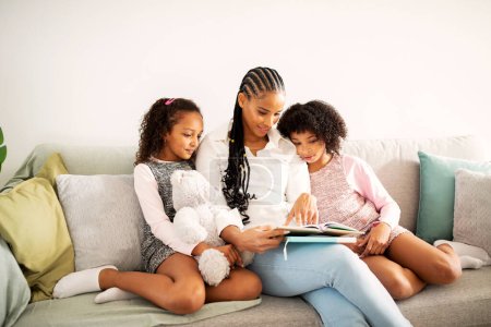 Photo for Black Mother And Two Little Daughters Reading A Book Sitting On Couch At Home. Happy Parent And Siblings Kids Spending Time Together On Weekend. Family Moments And Leisure Concept - Royalty Free Image