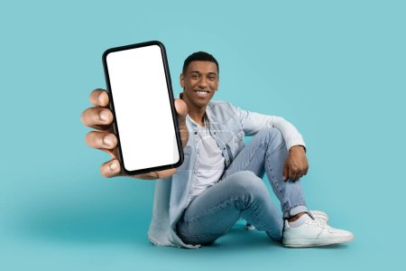 Photo for Cheerful millennial black male sitting on floor showing smartphone with empty screen isolated on blue studio background. Human emotion, recommendation app, device and blog - Royalty Free Image