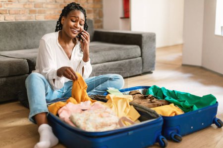Foto de Mujer negra feliz teniendo llamada telefónica mientras empaca la ropa en la bolsa de viaje, preparándose para el viaje de vacaciones, sentado en el suelo y hablando por teléfono celular, espacio libre - Imagen libre de derechos