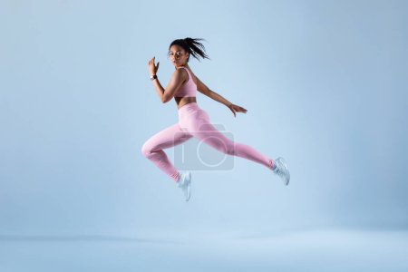 Photo for Training concept. Fit black woman in sportswear jumping or running, exercising on neon blue studio background, free space, full length. Energy and sport - Royalty Free Image