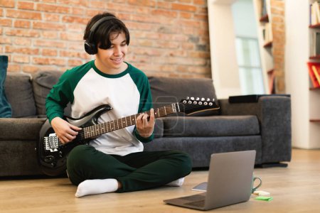 Photo for Smiling Asian Student Guy Playing Music On Electric Guitar Sitting Near Laptop On Floor At Home. Boy Learning Chords Online Via Computer On Weekend. Hobby And Leisure Concept. Selective Focus - Royalty Free Image