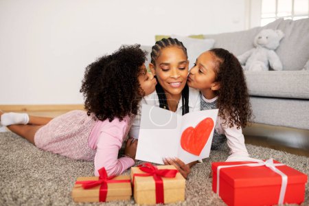 Téléchargez les photos : Fête des Mères. Filles noires heureuses embrassant maman alors qu'elle tient une carte de vœux avec un cœur peint, posant ensemble parmi des cadeaux enveloppés à la maison. Vacances en famille Concept - en image libre de droit