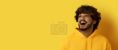 Foto de Emocionado emocionalmente sorprendido guapo rizado joven indio hombre en sudadera con capucha amarilla mirando espacio de copia y muecas sobre fondo de estudio amarillo, comprobar gran oferta o oferta, web-banner - Imagen libre de derechos