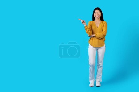 Photo for Full length studio shot of cheery beautiful millennial asian lady in nice outfit posing on blue background, pointing at copy space and smiling, young chinese woman showing nice offer, panorama - Royalty Free Image