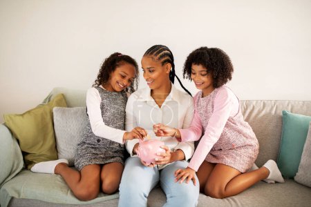 Photo for Smiling African American Mom And Her Daughters Raising Money Putting Coins In Piggybank Together Sitting On Couch Posing At Home. Great Bank Offer, Family Savings Safety Concept - Royalty Free Image