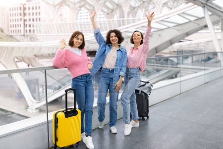 Foto de Retrato de tres amigas felices saludando con las manos en la cámara mientras posan en el aeropuerto, alegres mujeres jóvenes viajando juntas, de pie con maletas en la terminal mientras esperan la salida del vuelo - Imagen libre de derechos