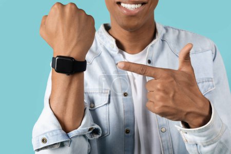 Photo for Cheerful millennial black male shows finger on fitness tracker or smart watch with blank screen, recommends offer, ad, app, isolated on blue studio background. Modern device for lifestyle - Royalty Free Image