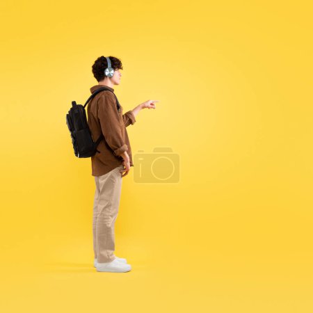 Téléchargez les photos : Vue latérale d'un étudiant portant un casque sans fil touchant un écran tactile invisible debout avec un sac à dos posant sur un fond jaune en studio. Carrée, pleine longueur - en image libre de droit