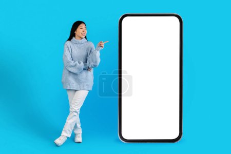 Photo for Cheerful pretty long-haired young asian woman in warm sweater and white pants posing on blue studio background, pointing at huge phone with empty screen mockup, showing nice offer, full length - Royalty Free Image