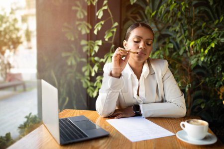 Photo for Thoughtful serious concentrated young black lady in suit writes on paper, thinks with laptop, enjoy coffee in cafe with green plants interior. Work, study and business, brainstorm and creative idea - Royalty Free Image
