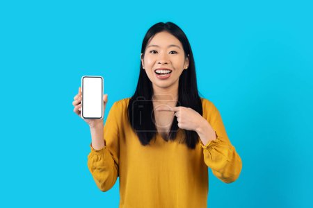 Photo for Modern smartphone with white empty screen in korean woman hand, cheerful pretty young asian lady showing great offer or newest mobile app, isolated on blue studio background, mockup - Royalty Free Image
