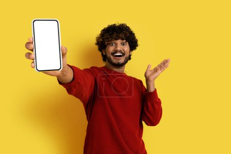 Foto de Emocional feliz guapo rizado milenario chico hindú en rojo mostrando teléfono celular moderno con pantalla blanca en blanco y gestos sobre fondo de estudio amarillo, maqueta, espacio libre para la publicidad - Imagen libre de derechos
