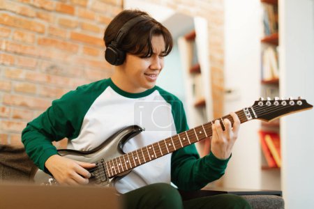 Foto de Asiático adolescente niño aprendiendo a tocar la guitarra eléctrica en línea sentado en el ordenador portátil en casa, usando auriculares inalámbricos. Concepto de Hobby para Adolescentes Modernos. Enfoque selectivo en Guy - Imagen libre de derechos