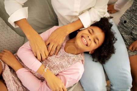 Photo for Cute Black Girl Laughing While Mother Tickling Her Having Fun Together At Home. Family Of Three Cuddling And Bonding On Weekend. Childhood Happiness Concept. Above View Shot - Royalty Free Image