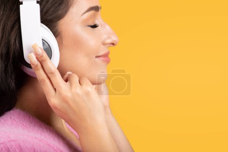 Téléchargez les photos : Happy relaxed jeune femme brune caucasienne dans des écouteurs sans fil avec les yeux fermés profiter de la musique, isolé sur fond de studio jaune, gros plan, profil. Repose-toi, écoute l'application audio seule - en image libre de droit