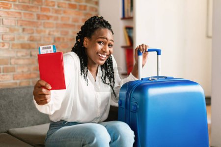 Foto de Listo para el viaje. Emocionada mujer afroamericana con pasaporte con boletos, sentada cerca de la maleta y sonriendo a la cámara en casa. Vacaciones, finalmente - Imagen libre de derechos