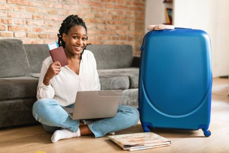 Téléchargez les photos : Femme afro-américaine positive montrant passeport et billet d'avion, en utilisant un ordinateur portable pour acheter des vacances touristiques à l'agence de voyage Internet, regardant et souriant à la caméra tout en étant assis valise soignée - en image libre de droit