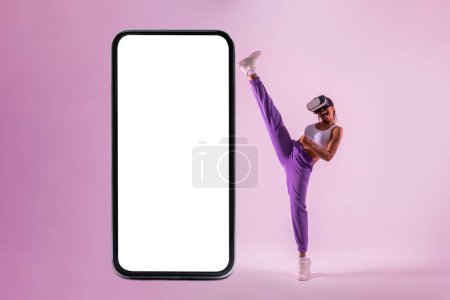 Photo for Fit african american lady in vr googles exploring virtual reality game near big smartphone with blank screen, mockup. Energetic black woman throwing kick - Royalty Free Image