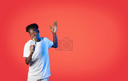 Foto de Guapo joven negro en blanco superestrella casual cantando con los ojos cerrados y el gesto, disfrutando de karaoke sobre fondo de estudio rojo en luz de neón, el uso de micrófono, espacio de copia - Imagen libre de derechos