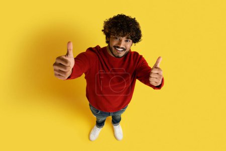 Photo for High angle view of happy handsome curly young indian man in stylish casual outfit showing thumb ups on yellow studio background, like something or someone, copy space. Human gestures concept - Royalty Free Image