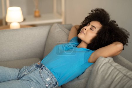 Foto de Sonriendo joven rizado mujer afroamericana con los ojos cerrados se encuentra en el sofá, disfruta de relajarse y dormir en el interior de la sala de estar. Dulces sueños, tarde de descanso, descanso en casa en tiempo libre - Imagen libre de derechos