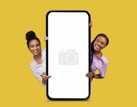 Téléchargez les photos : Offre en ligne. Deux belles femmes noires souriantes regardant derrière Big Blank Smartphone avec écran blanc isolé sur fond jaune, les femmes afro-américaines montrant l'espace de copie, collage, maquette - en image libre de droit
