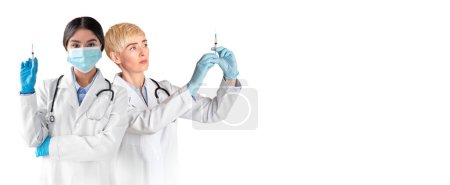 Photo for Two Female Doctors With Syringes In Hands Standing Isolated Over White Background, Prefessional Medical Workers In Uniform And Protective Masks Ready For Vaccination, Panorama With Free Space - Royalty Free Image