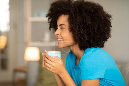 Photo for Happy young curly african american female with cup of hot drink, enjoy relaxing and aroma in living room interior, close up, profile. Coffee break, rest with tea at home in free time - Royalty Free Image