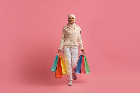 Téléchargez les photos : Belle jeune femme musulmane dans le hijab marchant avec des sacs à provisions lumineux sur fond rose studio, heureux Moyen-Orient femme portant foulard bénéficiant de ventes saisonnières et des réductions, Copier l'espace - en image libre de droit