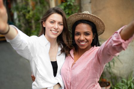 Photo for Glad young multiethnic ladies make selfie on phone, enjoy trip in new city, have video call, shooting video for travel blog outside. Fun together, vacation and summer, app for communication - Royalty Free Image