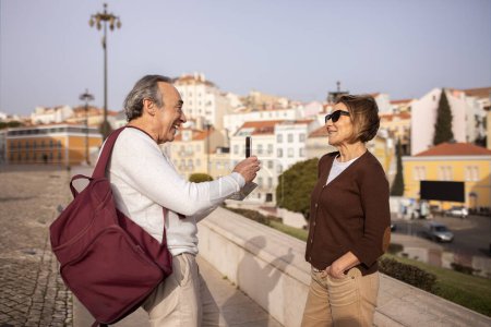 Téléchargez les photos : Des vacances. Heureux mari mûr prenant des photos de sa femme via Smartphone debout avec sac à dos à l'extérieur. Couple de personnes âgées européennes profitant de la vie en voyage à la retraite et s'amusant à Lisbonne - en image libre de droit