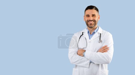 Foto de Médico profesional masculino en uniforme blanco con estetoscopio posando cruzando las manos y sonriendo, de pie sobre fondo azul, panorama, espacio libre. Concepto médico y de profesión médica - Imagen libre de derechos