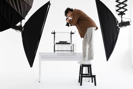 Téléchargez les photos : Photographe professionnel homme debout sur l'échelle et prendre des photos de contenu grand angle pour boutique moderne, travaillant au photostudio professionnel, vue de côté - en image libre de droit