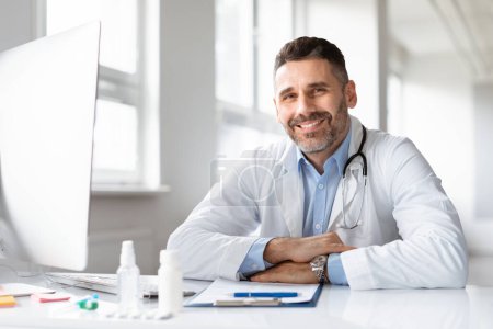 Foto de Retrato de un médico varón feliz vestido con uniforme médico blanco, sentado en el escritorio en el hospital y sonriendo a la cámara, espacio libre. Hombre GP o médico que trabaja en clínica privada - Imagen libre de derechos