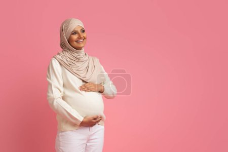 Foto de Mujer musulmana embarazada en hijab abrazando su vientre y mirando hacia otro lado, dama islámica esperando al bebé sonriendo y acariciando la barriga, expectante hembra del medio oriente posando sobre fondo rosa en el estudio - Imagen libre de derechos