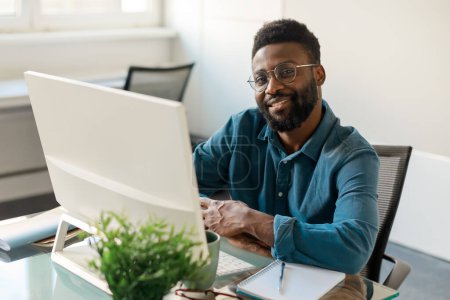 Téléchargez les photos : Homme d'affaires noir positif aux lunettes souriant à la caméra, assis devant l'ordinateur au bureau à l'intérieur du bureau, espace de copie. Carrière, chef d'entreprise - en image libre de droit