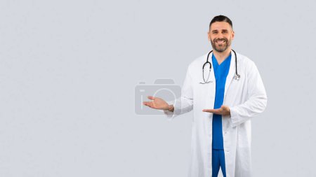 Téléchargez les photos : Publicité des soins de santé. Médecin masculin positif pointant de côté à l'espace libre avec deux mains, médecin d'âge moyen homme en uniforme debout sur fond gris, panorama avec place libre pour la conception - en image libre de droit