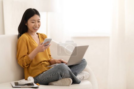 Téléchargez les photos : Dépendance au gadget. Positif joyeux jolie jeune femme japonaise pigiste travaillant de la maison, femme assise sur le canapé, en utilisant un ordinateur portable et en vérifiant les messages sur smartphone, copier l'espace - en image libre de droit