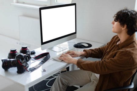 Téléchargez les photos : Graphiste masculin ou photographe travaillant avec un ordinateur à écran vide dans son espace de travail, assis au bureau avec un appareil photo et un équipement d'objectif, éditant des photos après la séance photo - en image libre de droit