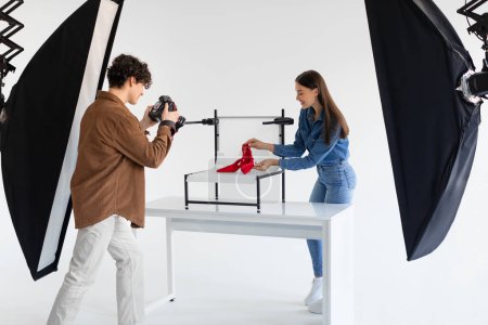 Photo for Young male photographer taking photo of stylish red shoes on white platform background in photostudio with modern equipment, lady helping to make content photoshoot - Royalty Free Image