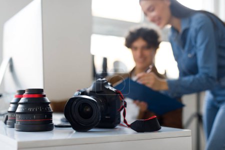 Photo for Male photographer and his assistant working together at workplace, discussing photoshoot, focus on professional photo camera and lens lying on table - Royalty Free Image