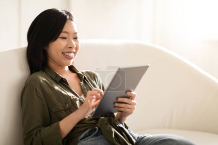 Photo for Relaxed cheerful young pretty asian woman in casual outfit sitting on white couch at home, reading blog on Internet, scrolling, chatting online, using digital tablet, copy space, closeup - Royalty Free Image