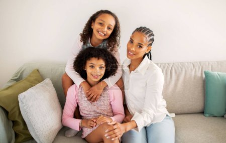 Téléchargez les photos : Joyeux afro-américain mère et filles étreignant collage dans le salon moderne assis sur le canapé à l'intérieur, souriant à la caméra. Tourné d'une famille heureuse de trois. Concept de maternité - en image libre de droit