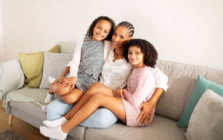 Foto de Feliz madre negra e hijas de niños abrazándose sentados en el sofá en casa, sonriendo mirando a la cámara. Shot de familia de tres vinculación en sala de estar moderna en fin de semana - Imagen libre de derechos