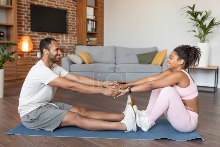 Photo for Cheerful young black couple in sportswear do muscle exercises for legs on mat, floor together in living room interior, profile. Active slimming and fitness at home, body care and workout due covid-19 - Royalty Free Image