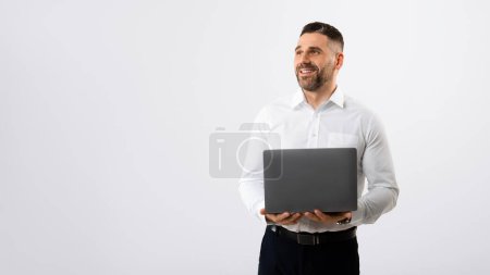 Téléchargez les photos : Employé prospère homme d'affaires en chemise blanche travaillant avec ordinateur portable PC et regardant de côté l'espace libre, debout sur fond de studio gris clair, panorama - en image libre de droit