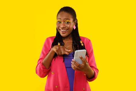 Téléchargez les photos : Joyeuse femelle noire pointant sur le téléphone portable recommandant application mobile souriant à la caméra posant debout en studio sur fond jaune. Lady publicité nouvelle application pour Smartphone - en image libre de droit