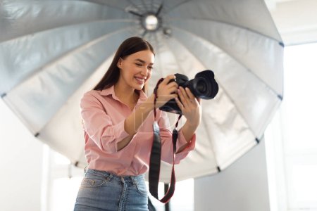 Photo for Young female photographer standing in front of reflective umbrella, working with professional dslr camera in modern photostudio, free space - Royalty Free Image